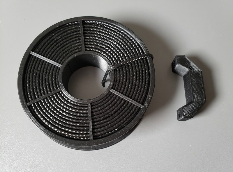 Spool of black carbon fiber composite 3D printer filament with visible fiber texture next to a printed black part on a workbench