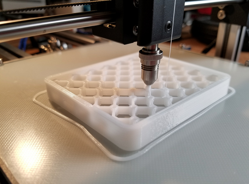 3D printer mid-print depositing a white PETG sensor enclosure with visible perimeter walls and infill pattern, build plate at slight angle, professional photography