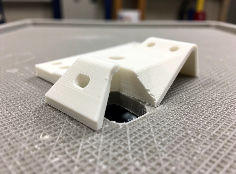 A 3D printed PLA bracket with a warped corner lifting off a textured print bed