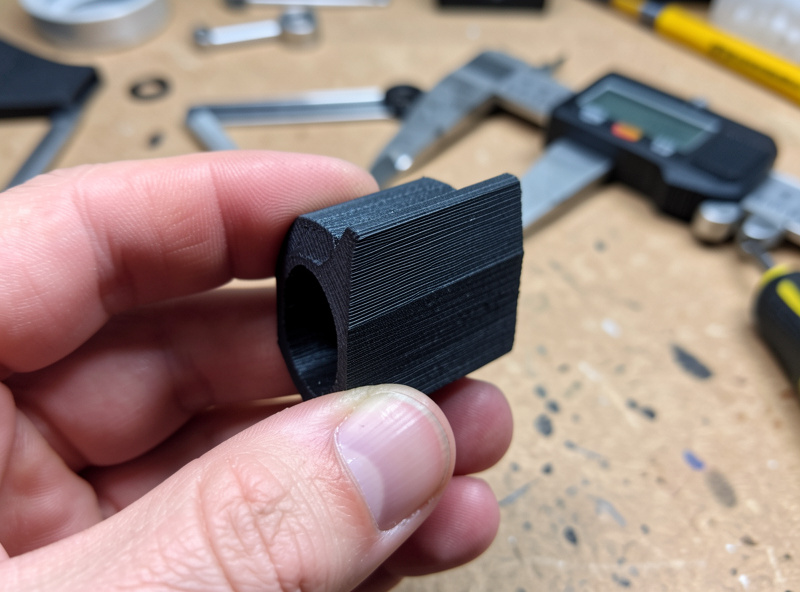 Hand holding a freshly 3D-printed black PETG bracket adapter showing layer lines, calipers nearby